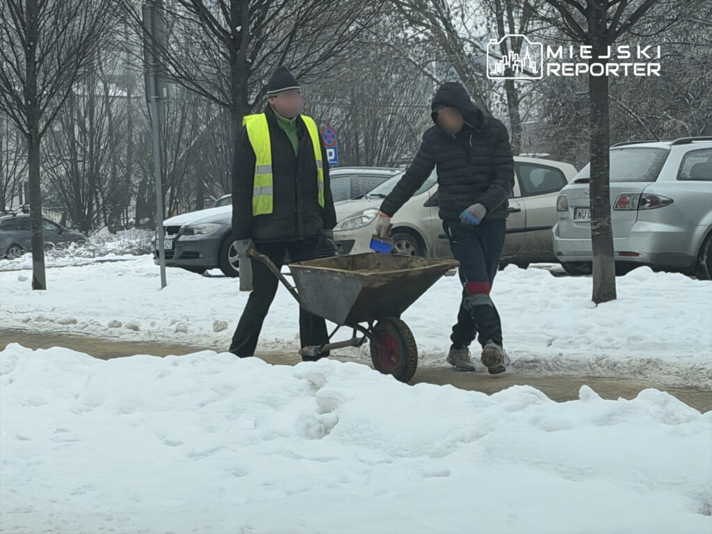 Mężczyzna w kamizelce odblaskowej i mężczyzna w zimowej kurtce transportują materiał budowlany na wózku w zaśnieżonym otoczeniu.