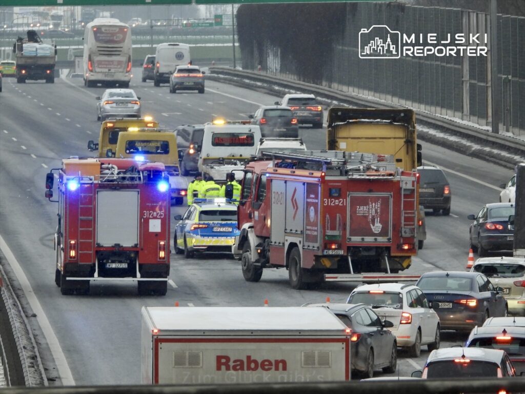 Na drodze zablokowanej przez wypadek, straż pożarna i policja zabezpieczają miejsce zdarzenia, a samochody stoją w korku.