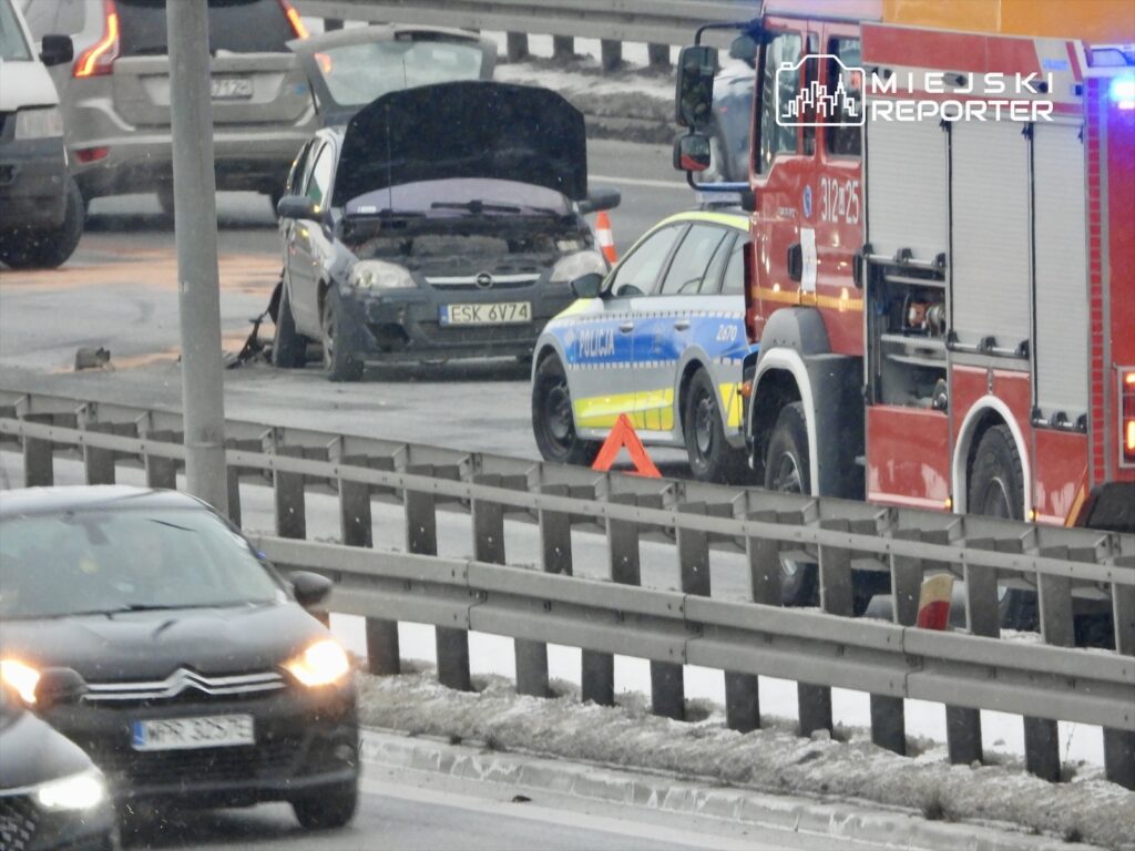 Na drodze stoi uszkodzony samochód z otwartą maską, obok policja i wóz strażacki. Ruch odbywa się wolno.