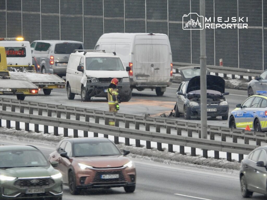 Strażak w kasku i mundurze interweniuje przy rozbitym samochodzie na zatłoczonej drodze, obok policyjny radiowóz.