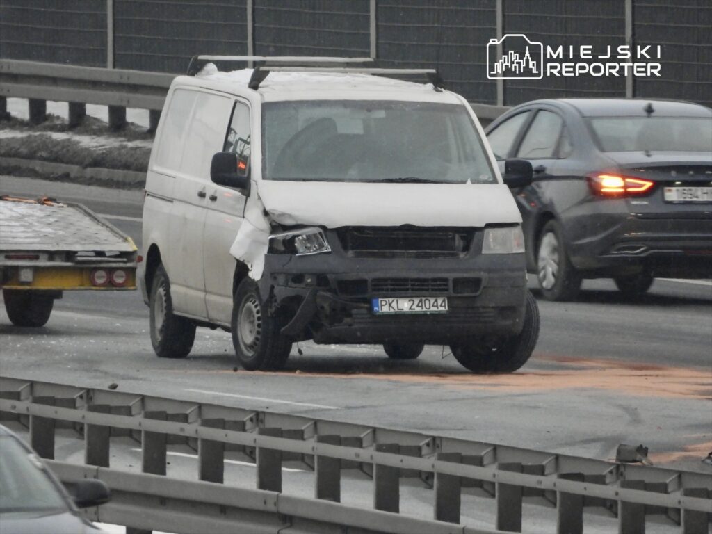 Biały furgon z uszkodzoną przednią częścią stoi na ruchliwej drodze, obok niego znajduje się laweta.