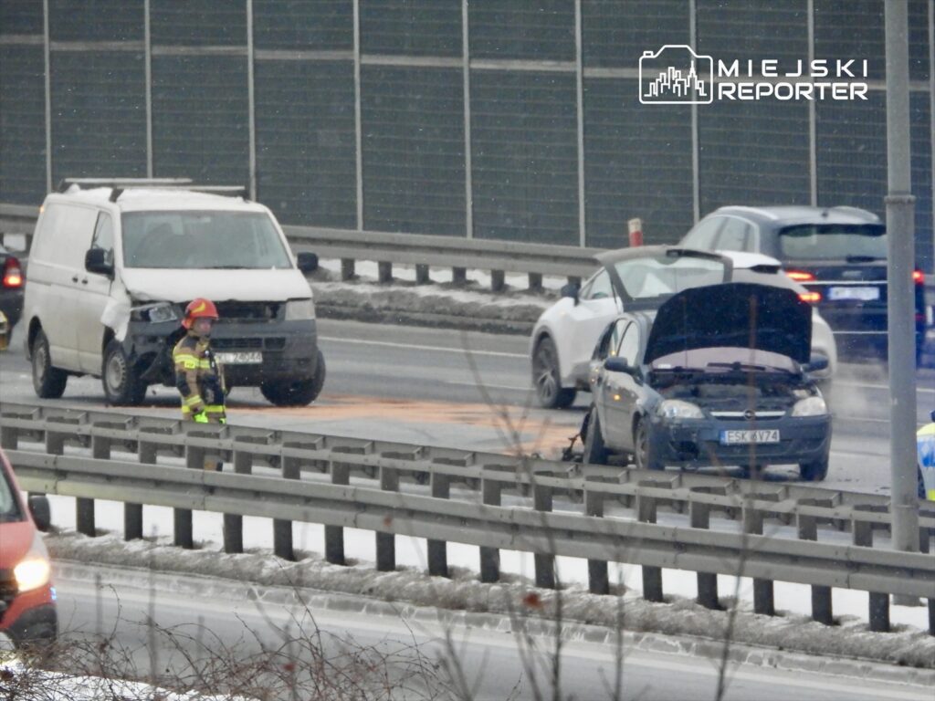 Strażak w kasku obserwuje wypadek na drodze, obok uszkodzonego samochodu osobowego i białego vana.
