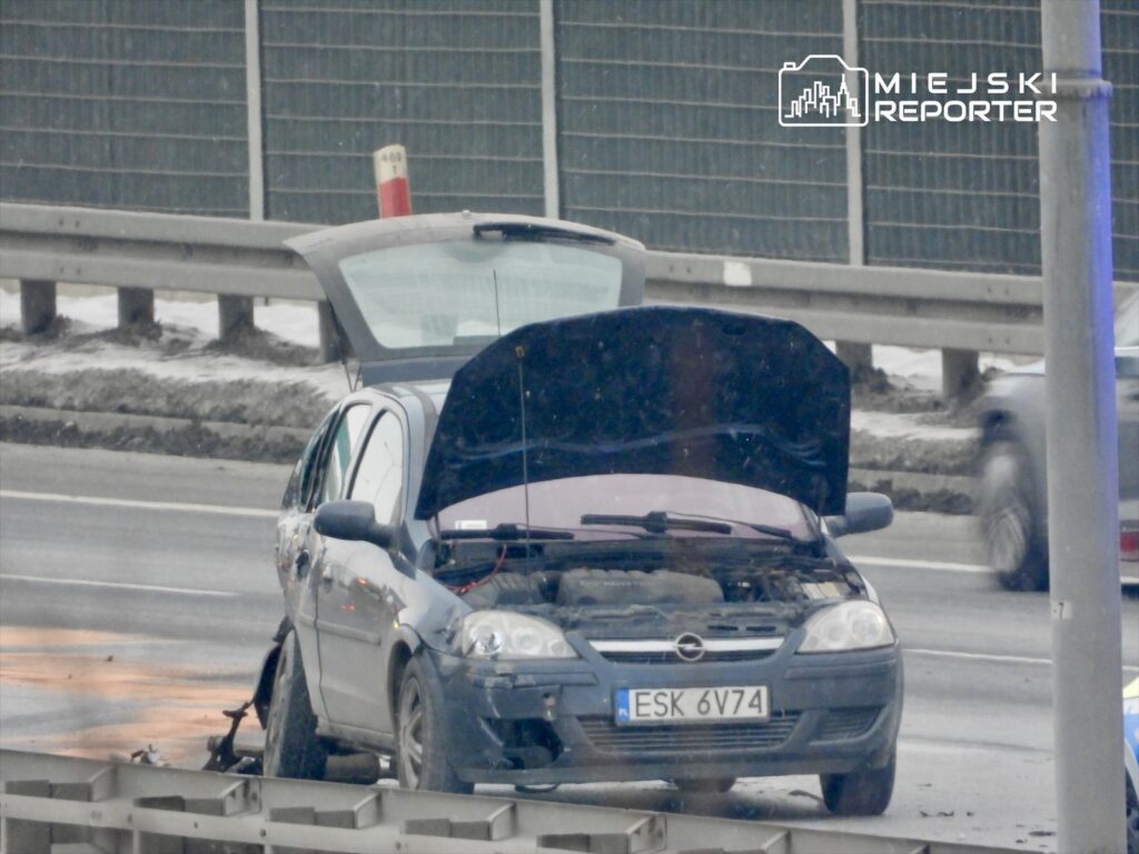 Uszkodzony niebieski samochód osobowy stoi na poboczu drogi z otwartą maską, w tle widać inne pojazdy.