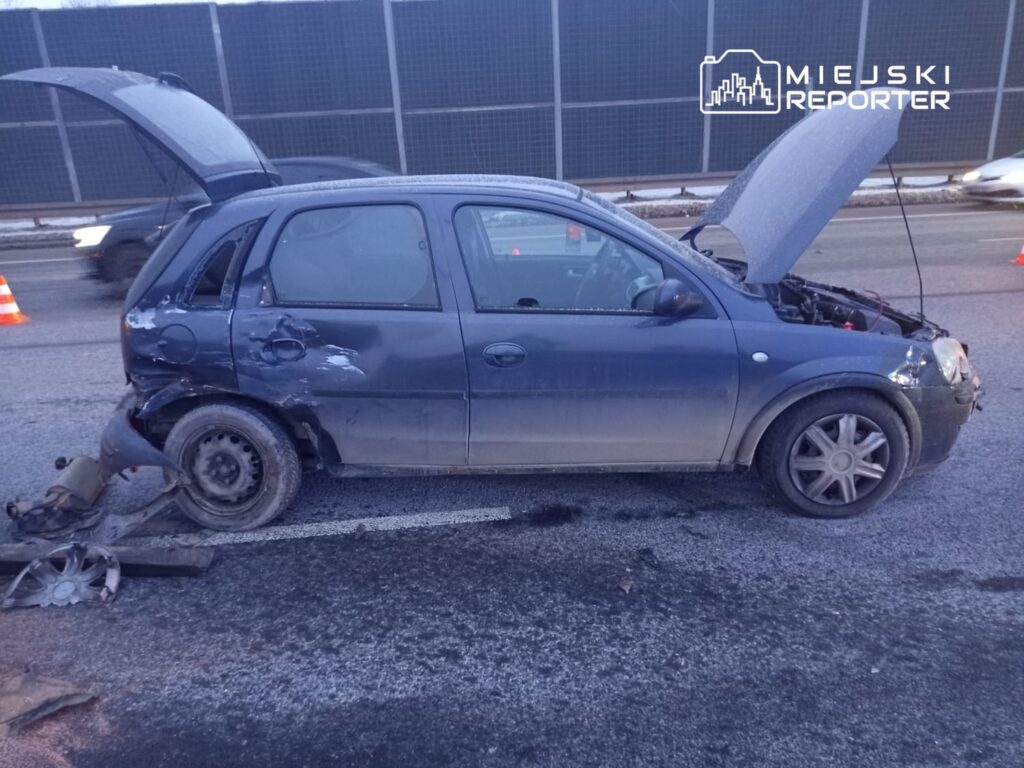 Uszkodzony niebieski samochód osobowy stoi na drodze, z otwartą maską i uszkodzeniami bocznymi.