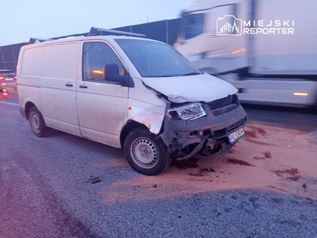 Uszkodzony biały samochód dostawczy stoi na drodze, obok widać rozlane płyny i znaki ostrzegawcze.