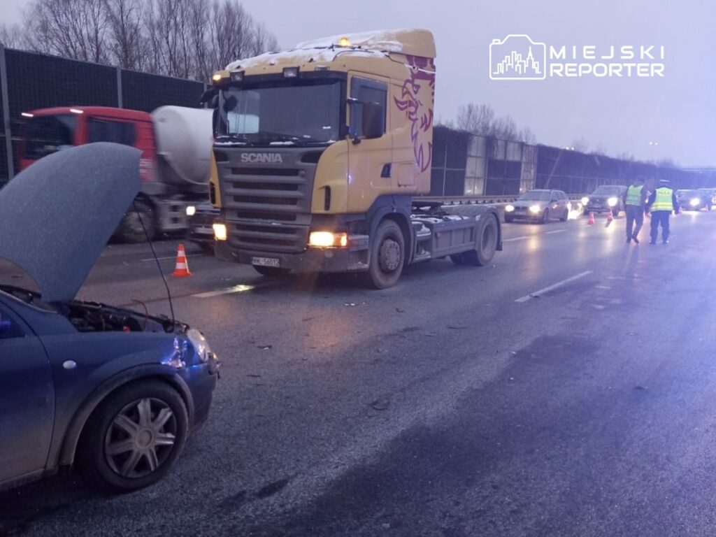 Czarny samochód osobowy z otwartą maską stoi na drodze obok dużego ciężarowego pojazdu, w tle widać inne samochody.