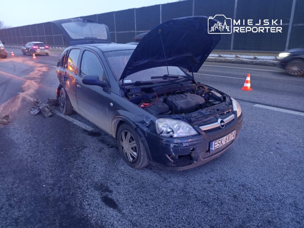Uszkodzony samochód osobowy stojący na poboczu drogi, z otwartą maską i uszkodzeniami nadwozia. W tle widoczne inne pojazdy oraz znaki ostrzegawcze.