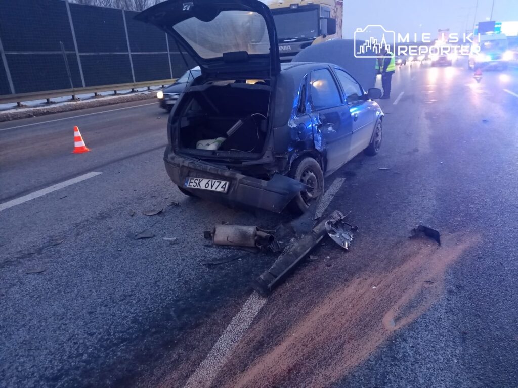 Uszkodzony samochód osobowy z otwartą klapą bagażnika leży na pasie drogowym, obok niego leżą fragmenty karoserii.