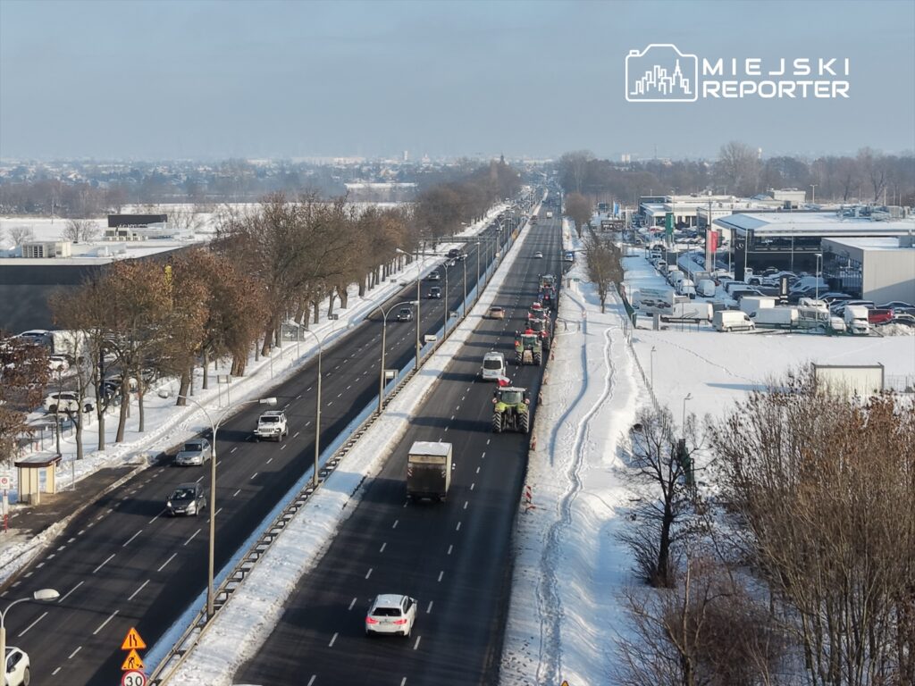 Ruch drogowy na ośnieżonej ulicy, z ciągnikami i samochodami w kolejce, w tle nowoczesne budynki.