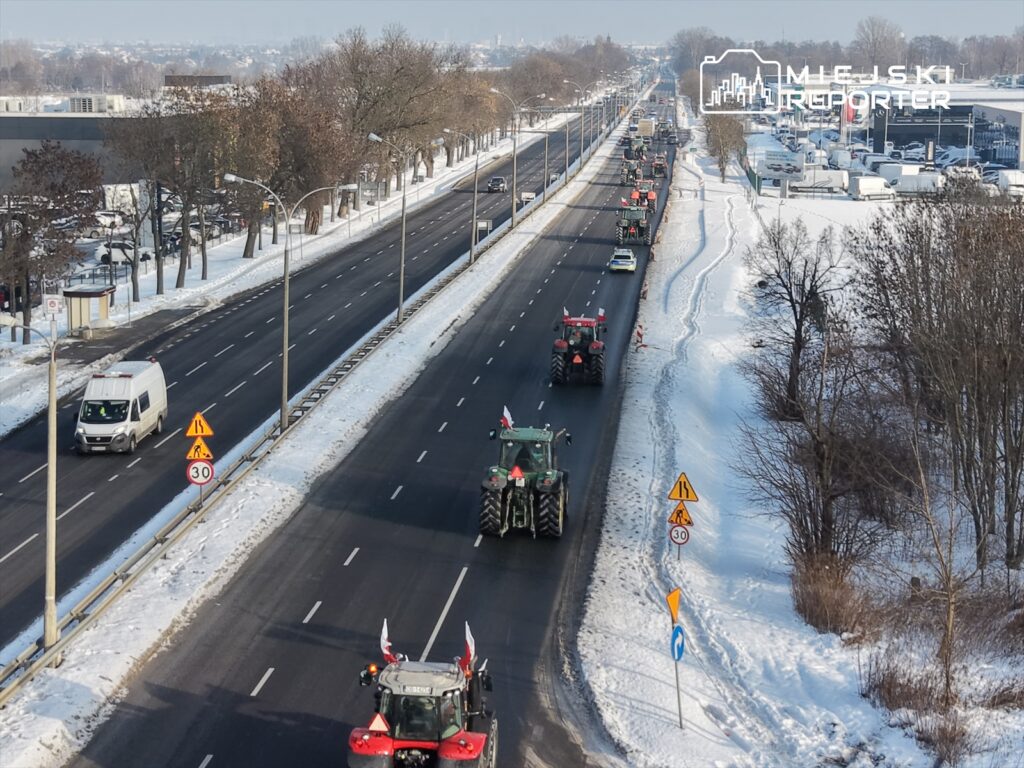 Traktory z flagami narodowymi przejeżdżają po zaśnieżonej drodze, obok drzew i znaków drogowych.