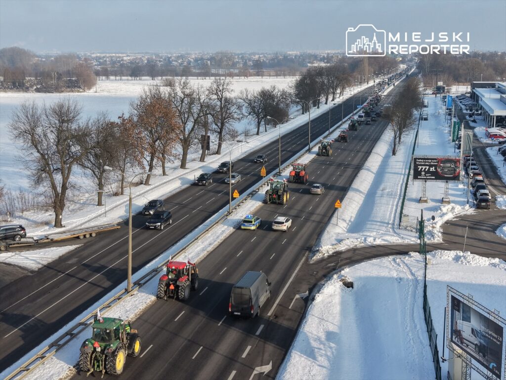 Traktory poruszające się wzdłuż ruchliwej drogi w zimowej scenerii, obok pokrytej śniegiem ziemi i drzew.