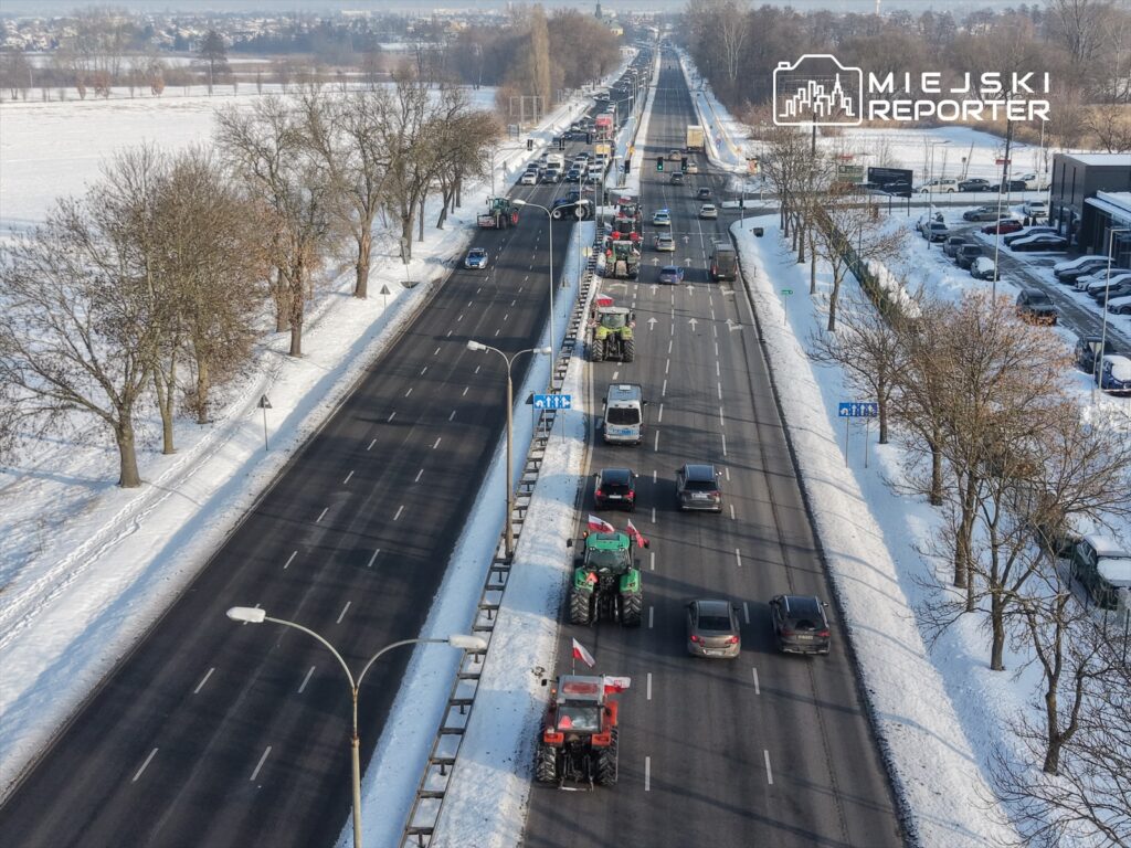 Traktory z flagami narodowymi w ruchu na zaśnieżonej drodze, obok stojące samochody osobowe i drzewa w tle.