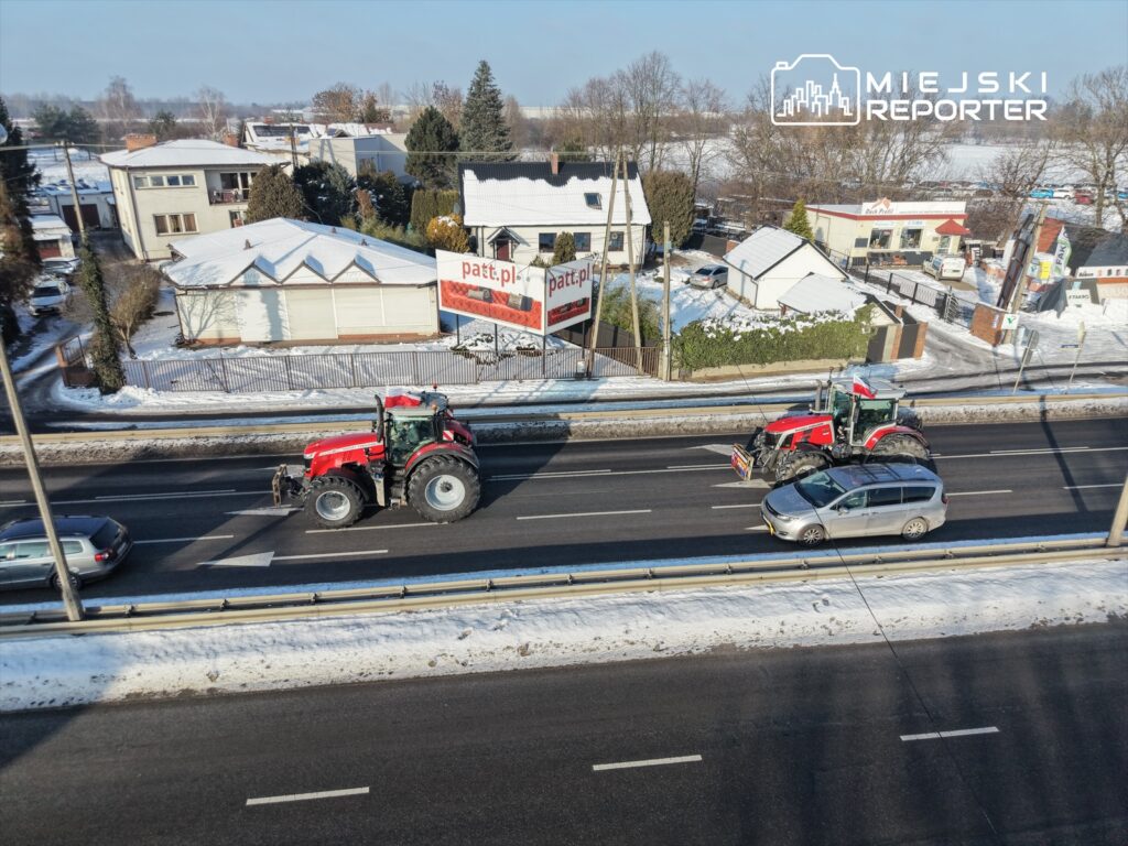 Czerwony traktor z pługiem jedzie po drodze, obok niego przejeżdża szary samochód osobowy, w tle widoczne domy.
