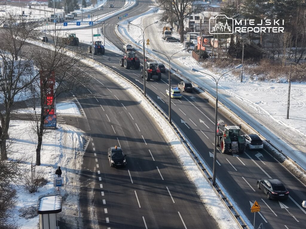 Traktory przemieszczają się po drodze w zimowym otoczeniu, obok samochodów osobowych i znaku drogowym.