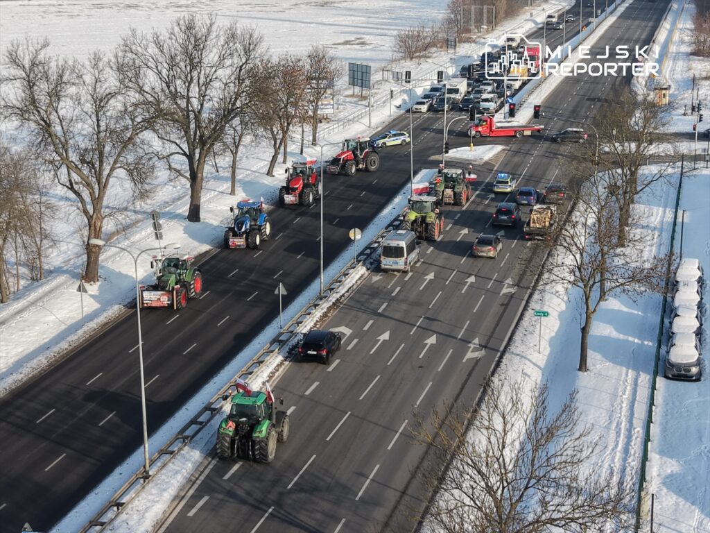 Traktory w kolumnie przejeżdżają przez ruchliwą drogę, obok zasypanych śniegiem drzew.