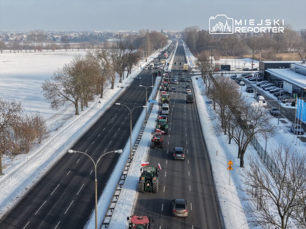 Ruch drogowy na szerokiej ulicy pokrytej śniegiem, z ciągnącymi się wzdłuż traktorami i samochodami osobowymi.