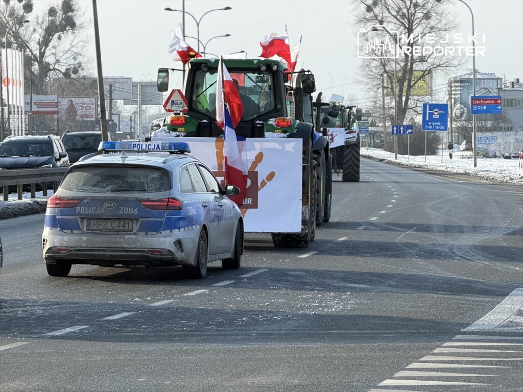 Traktor z transparentem i flagami narodowymi jedzie ulicą, a obok znajduje się radiowóz policji.