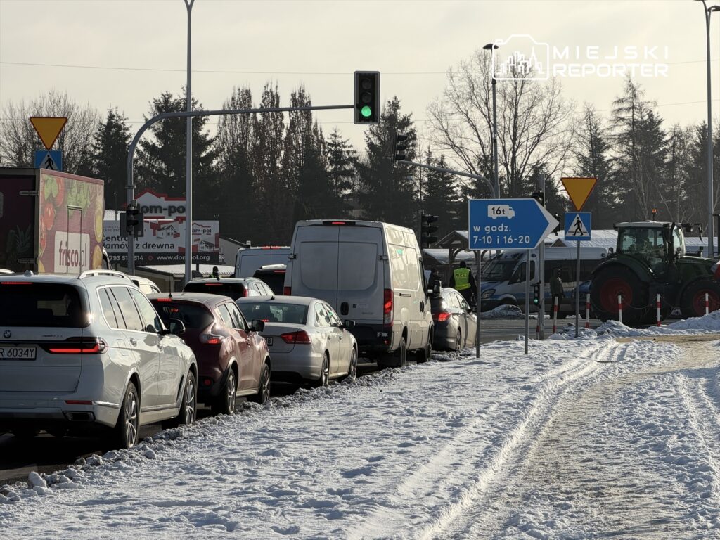 Korki samochodowe na drodze w zimowym otoczeniu, z sygnalizacją świetlną i oznakowaniem drogowym.