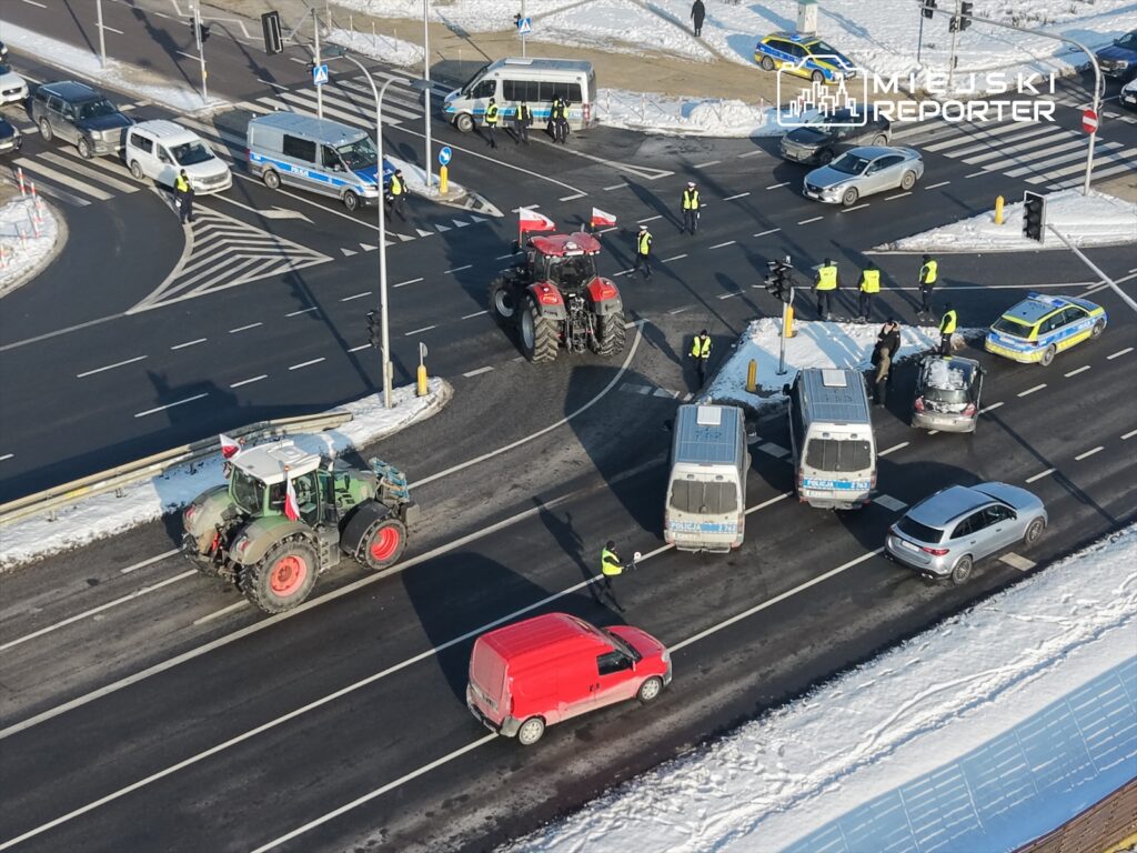 Traktor z flagą narodową przejeżdża przez rondo, a policjanci kierują ruchem wokół zablokowanych pojazdów.