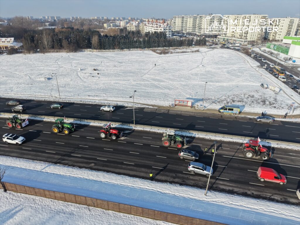 Traktory poruszające się po ruchliwej drodze w zimowej scenerii, obok samochodów osobowych.