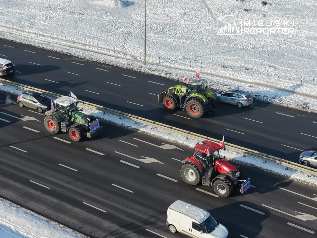 Traktory z flagami zatrzymane na ruchliwej drodze, obok samochodów osobowych, w zimowym otoczeniu.