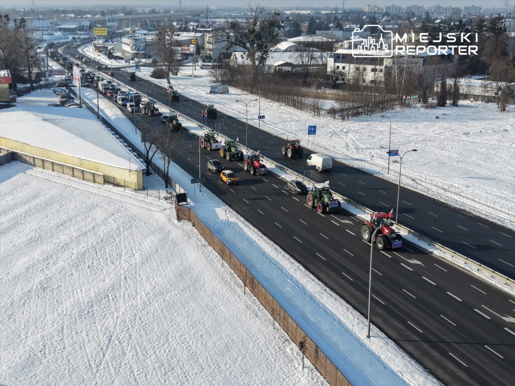Traktory z flagami narodowymi poruszają się w kolumnie po ośnieżonej drodze w miejskim otoczeniu.