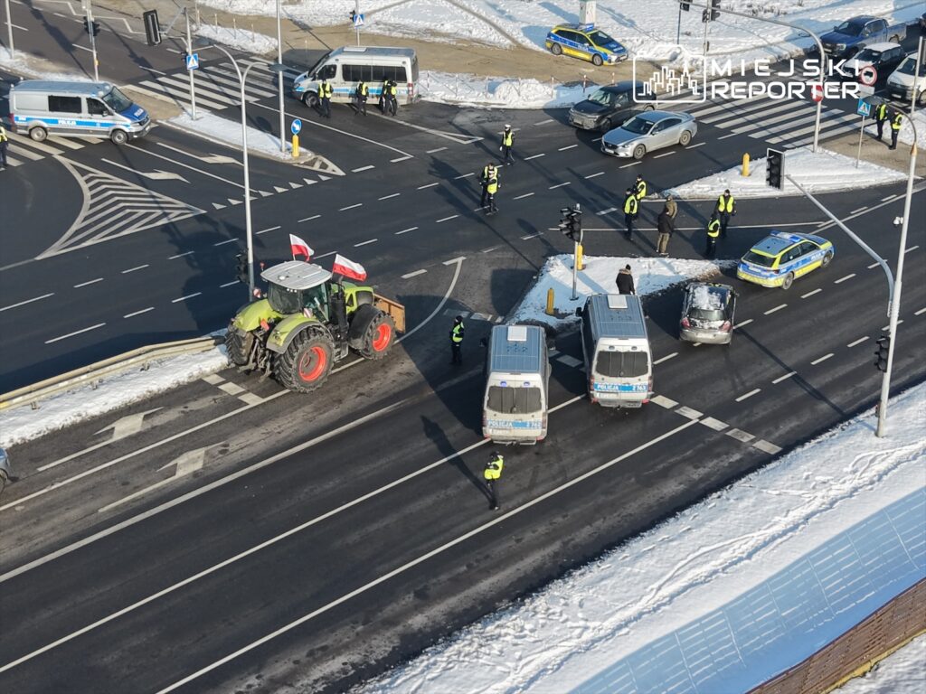 Traktor z polską flagą blokuje ruch na skrzyżowaniu, obok stoją policyjne radiowozy i funkcjonariusze w kamizelkach.