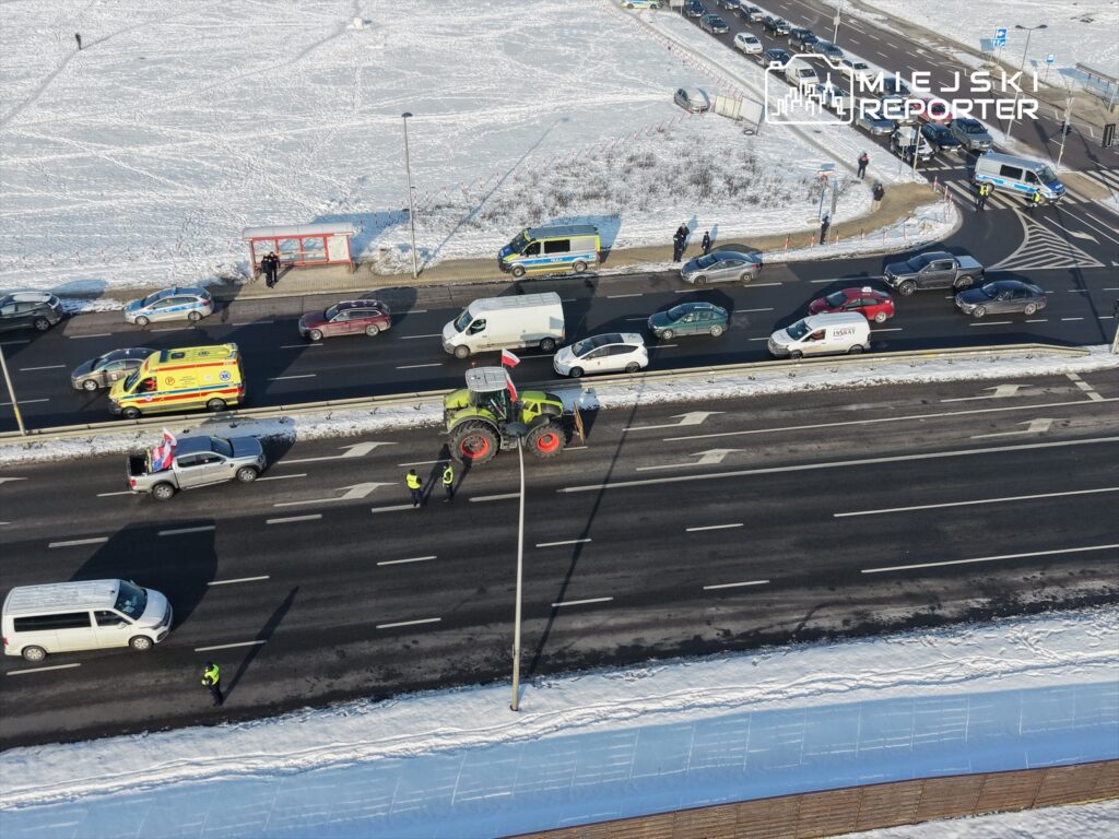 Traktor z flagą narodową zatrzymany na ruchliwej drodze, obok policjanci kierujący ruchem w zimowej scenerii.