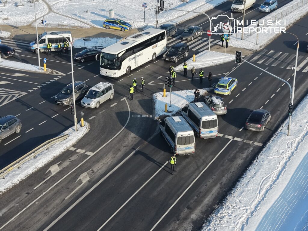 Grupa policjantów kieruje ruchem na skrzyżowaniu, obok stoi autobus i kilka samochodów osobowych. W tle widać zaspy śniegu.