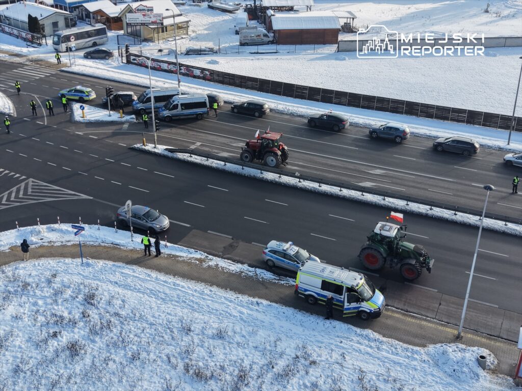 Traktor z polską flagą przejeżdża obok radiowozu, a policjanci kierują ruchem na zaśnieżonej drodze.