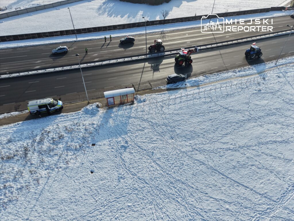 Traktory przejeżdżają obok zatrzymanych samochodów na zaśnieżonej drodze, obok stoi radiowóz.