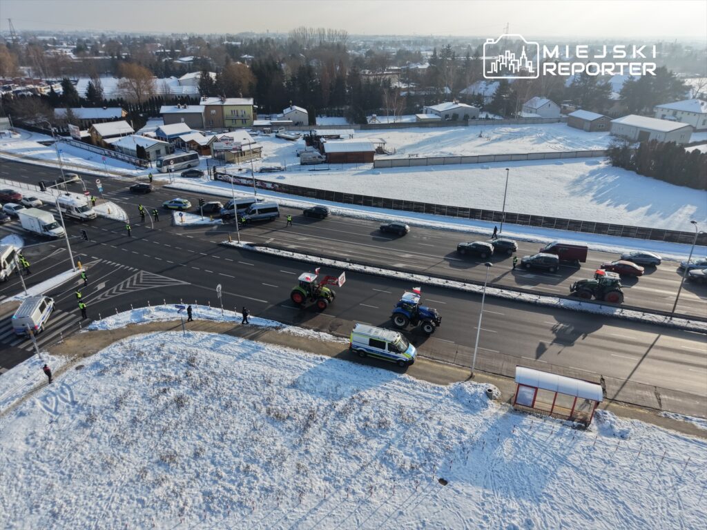Traktory z flagami przejeżdżają obok stojących samochodów na zaśnieżonej drodze, w otoczeniu policji i obserwujących ludzi.