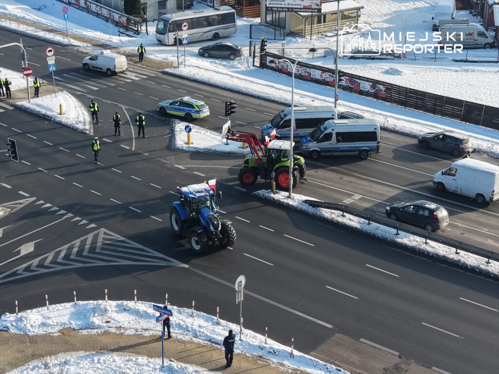 Traktor z flagą narodową przejeżdża przez skrzyżowanie, a policjanci kontrolują ruch w zaśnieżonym otoczeniu.