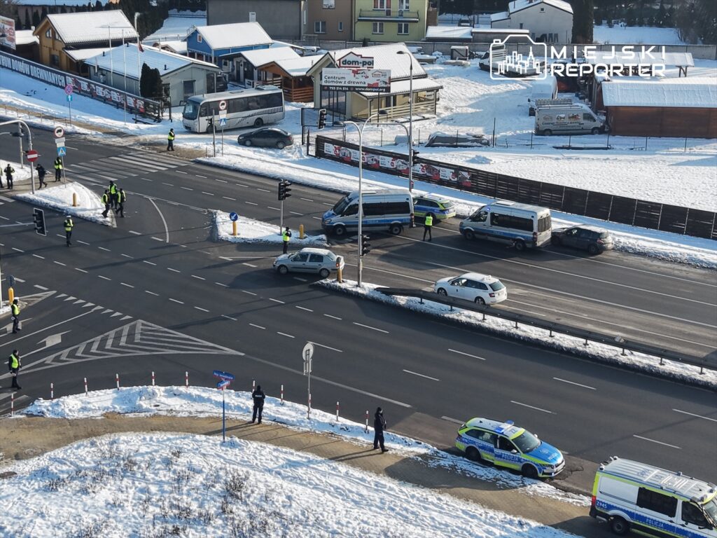 Grupa policjantów w odblaskowych kamizelkach kieruje ruchem na skrzyżowaniu, obok parkowane pojazdy i śnieżna nawierzchnia.