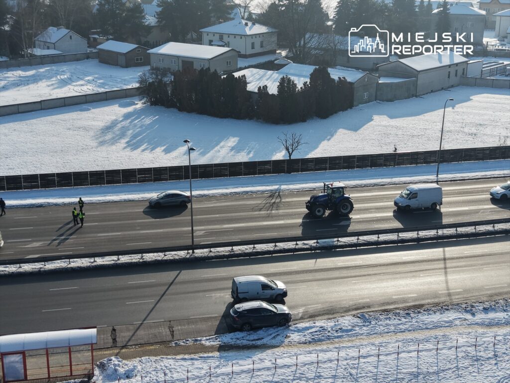 Traktor porusza się po drodze, obok policjanci rozmawiają z kierowcami, w tle śnieżne pole i domy.