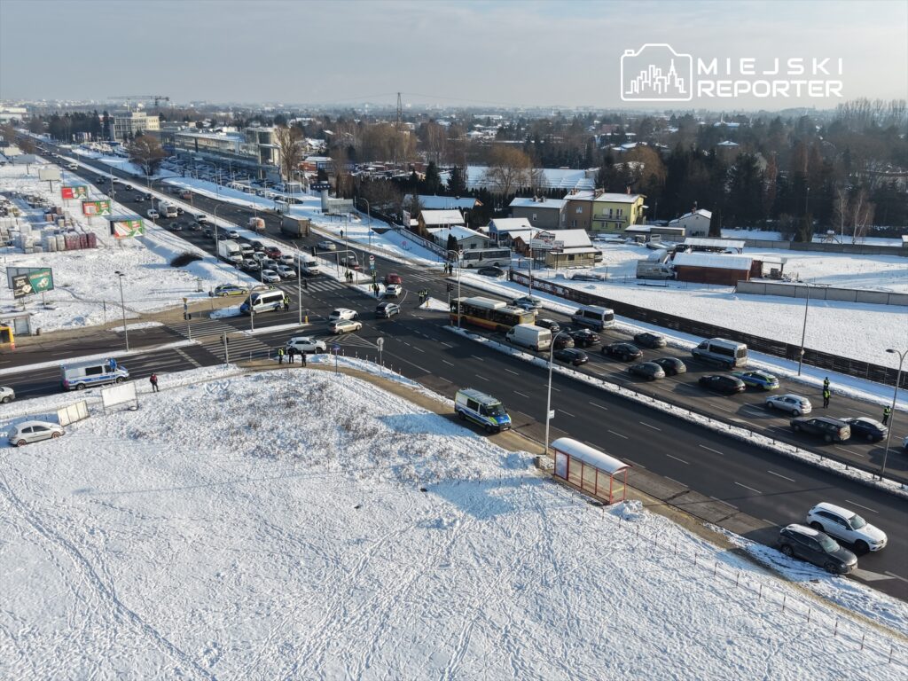 Ruch samochodowy na skrzyżowaniu w zimowej scenerii, z policją kontrolującą pojazdy w tle.