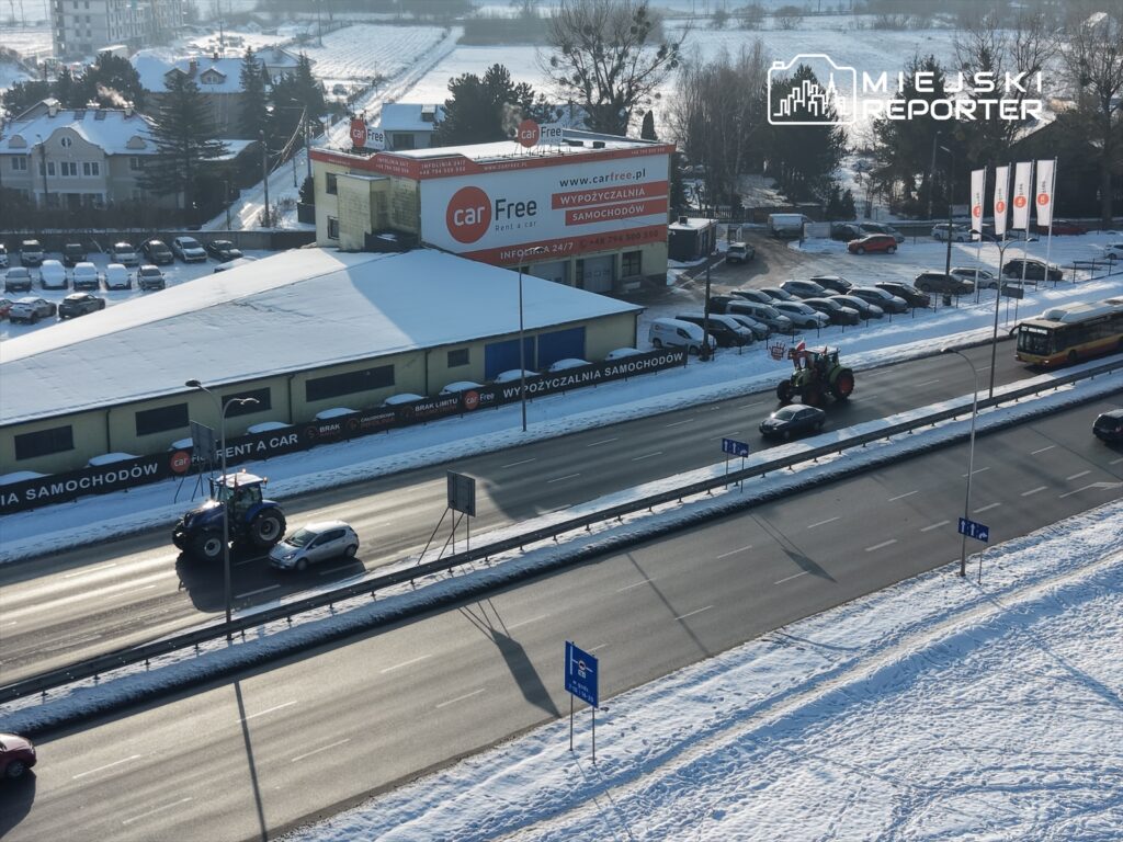 Traktor porusza się po ośnieżonej drodze obok wypożyczalni samochodów, a w tle widać samochody zaparkowane na parkingu.