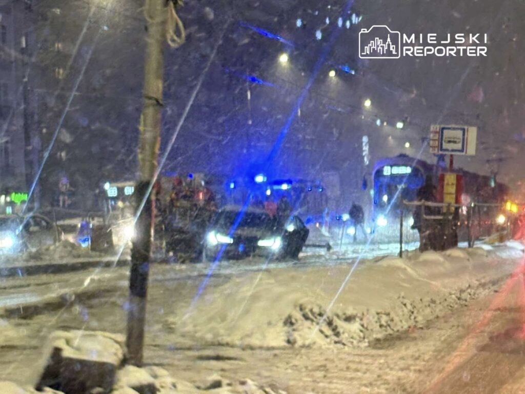 Policyjne samochody z włączonymi sygnałami świetlnymi stoją na zaśnieżonej ulicy w nocy, obok tramwaju.