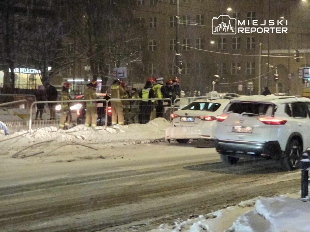 Grupa strażaków w kaskach rozmawia z policjantami przy ogrodzeniu w zaśnieżonej ulicy.