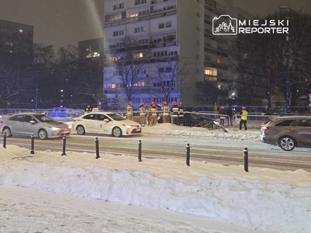 Grupa ratowników w odblaskowych kamizelkach pracuje przy zablokowanej drodze w zimowej scenerii, obok zaparkowanych samochodów.