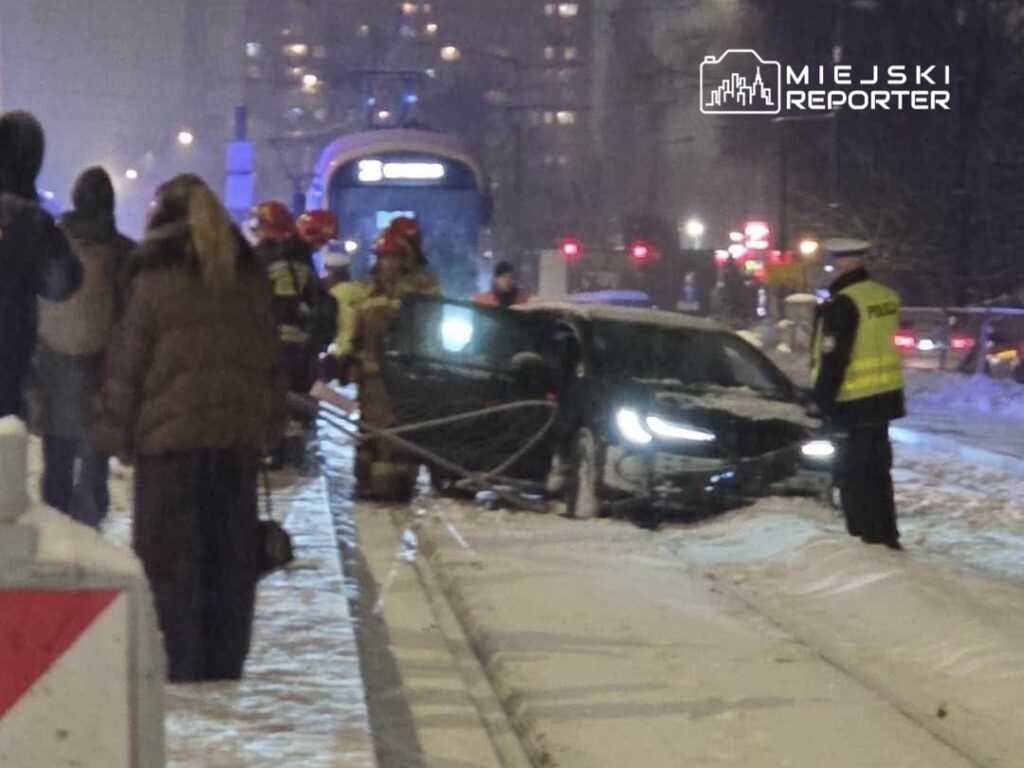 Grupa osób obserwuje akcję ratunkową, podczas gdy strażacy pomagają wydostać samochód z zaspy śnieżnej w miejskim otoczeniu.