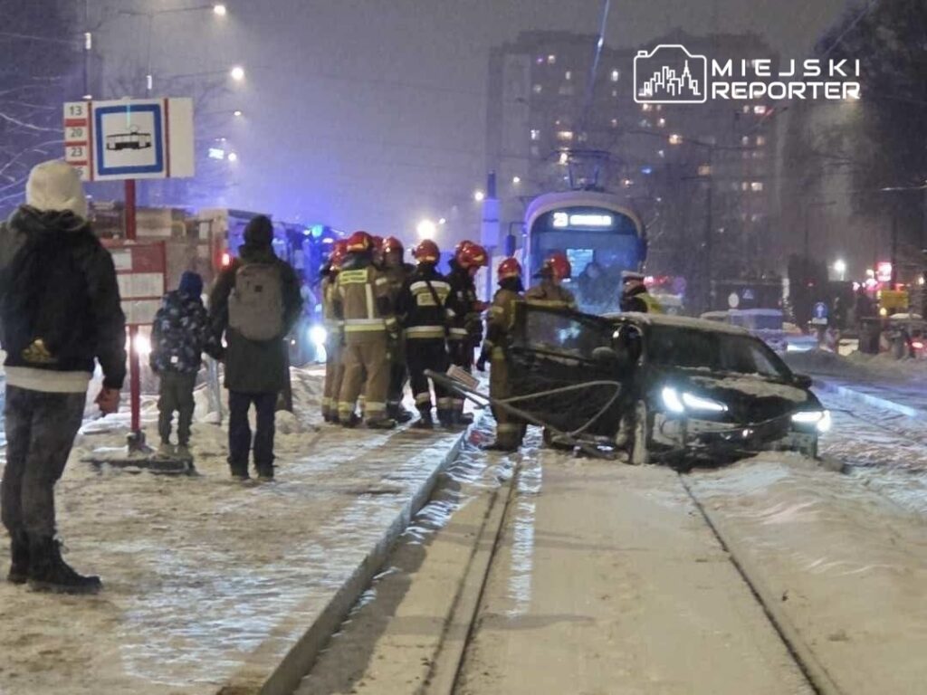 Grupa strażaków w mundurach, pracująca przy wypadku samochodowym na ośnieżonej ulicy, obok tramwaju w nocy.