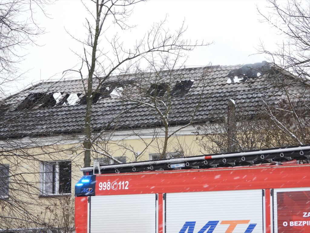 Zniszczony dach budynku z widocznymi otworami po pożarze, obok wóz strażacki z włączonymi sygnalizatorami.