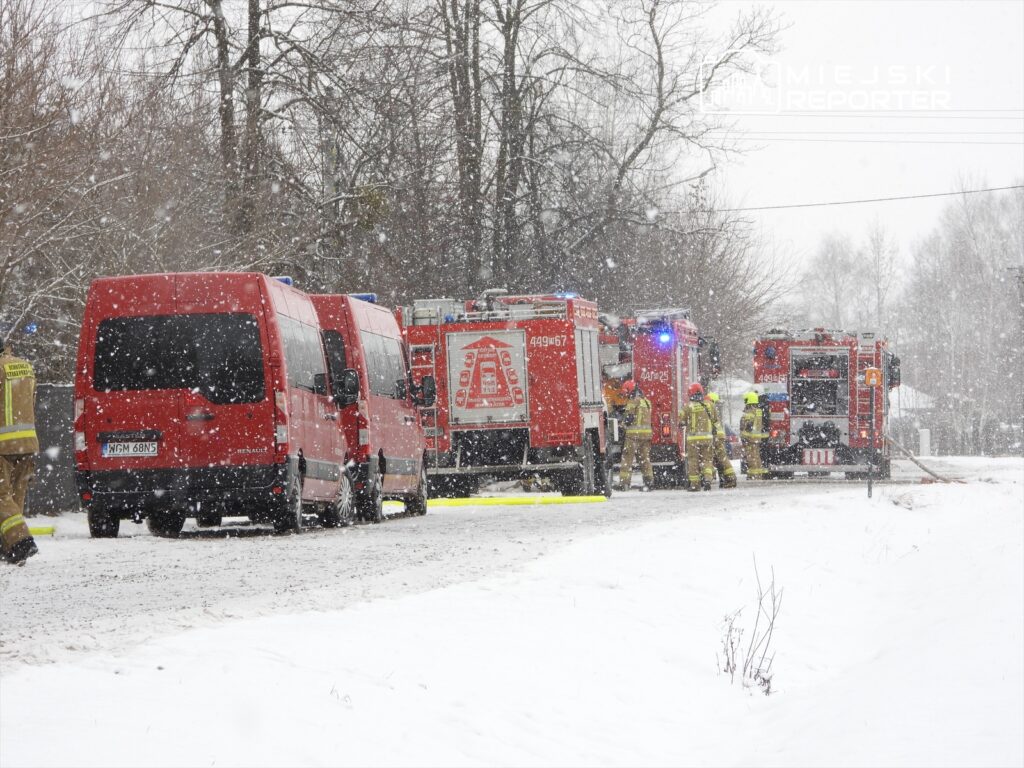 Czerwone pojazdy strażackie zaparkowane na zaśnieżonej drodze, ratownicy w mundurach przygotowujący sprzęt.