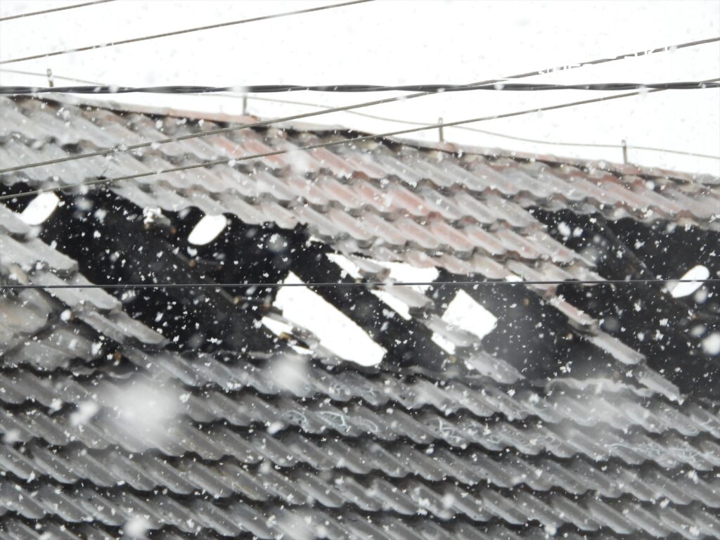 Uszkodzony dach z widocznymi dziurami, przez które pada śnieg, w tle szare niebo.
