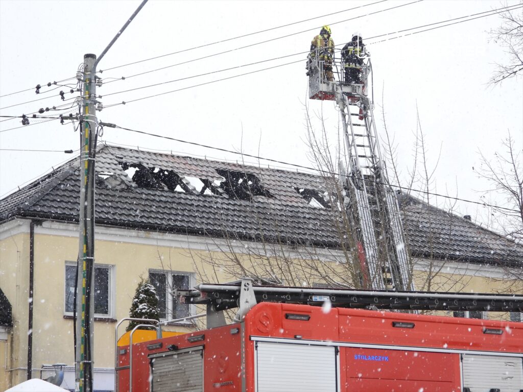 Strażacy na podnośniku gaszą pożar dachu budynku w trakcie opadów śniegu. W tle widoczna czerwona strażacka ciężarówka.