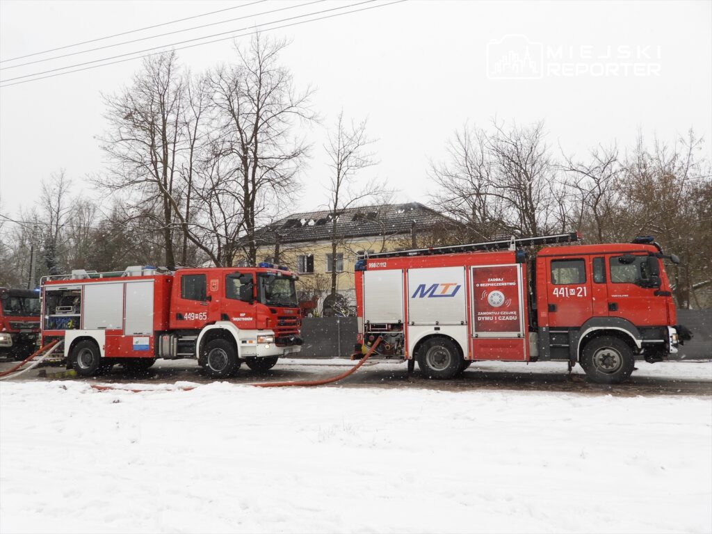 Dwa wozy strażackie z wężami naśnieżonej ulicy obok budynku mieszkalnego w zimowej scenerii.