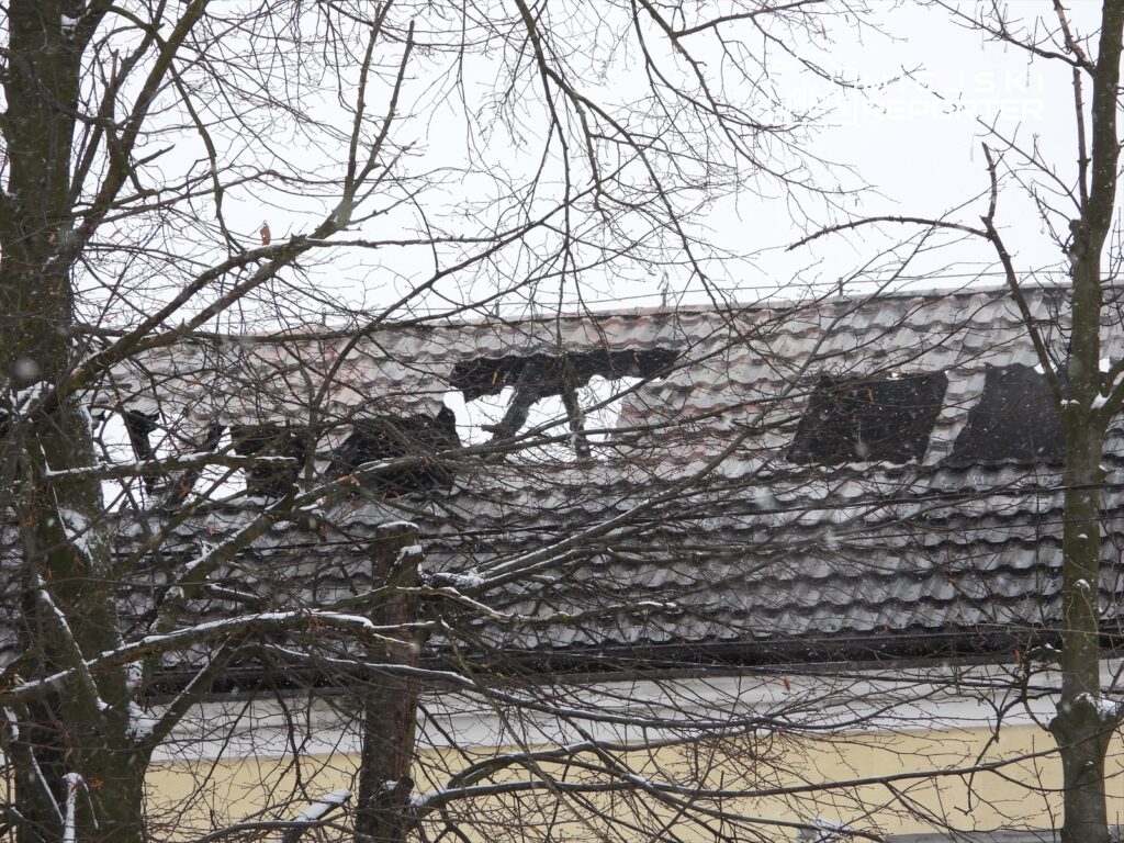 Zniszczony dach budynku z lukami, widoczny przez gałęzie drzew pokrytych śniegiem.