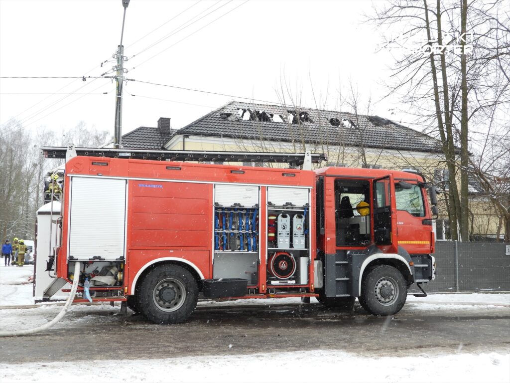 Czerwony wóz strażacki z wężem na drodze, w tle budynek z uszkodzonym dachem, pada śnieg.