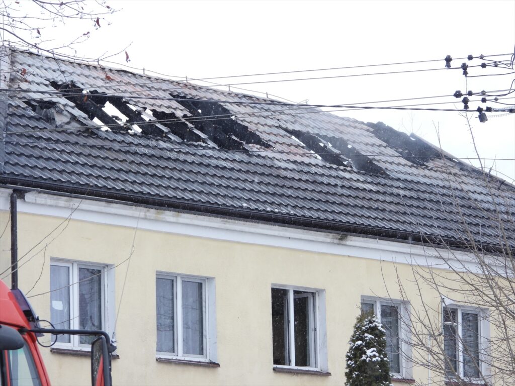 Uszkodzony dach budynku mieszkalnego z widocznymi dziurami i uszkodzeniami, w tle gołe gałęzie drzew.